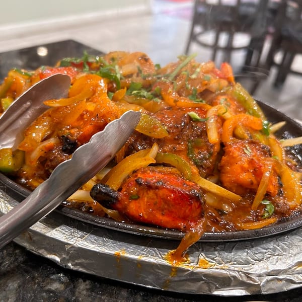 Sizzling Indian chicken tikka with onions and peppers on a hot platter, served with tongs.
